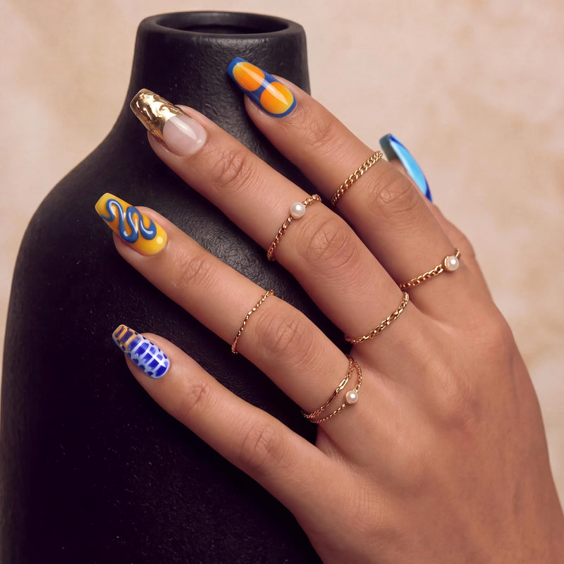 Lifestyle hand model wearing Solar Tide press-on nails with blue and orange wave designs, metallic gold accent nails, and gold rings