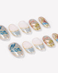 Angled flat lay of Stained Glass Leaves press-on nails with translucent blue, green, pink, and cream panels, raised gold leaf outlines, and shimmer finish