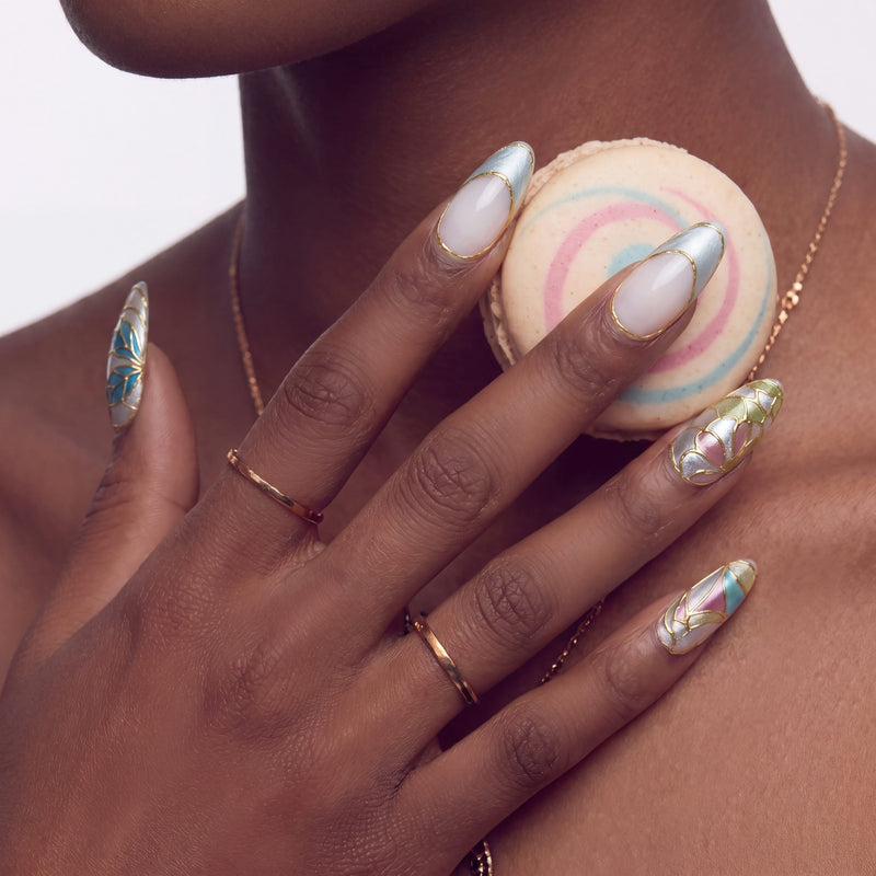 Lifestyle hand model wearing Stained Glass Leaves press-on nails with gold outlined leaf panels, blue and cream tips, and a pastel macaron prop