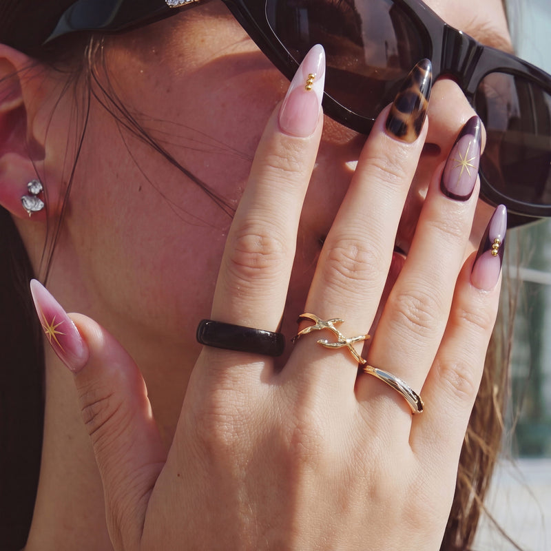 Close-up hand model wearing Starlit Mocha press-on nails with pink shimmer, gold starbursts, black mocha accents, and gold rings