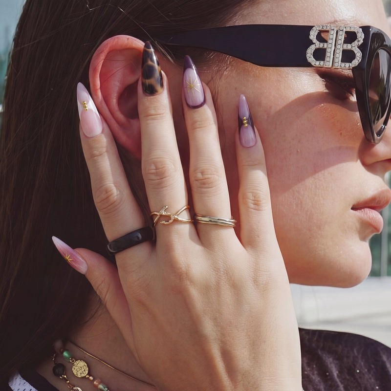 Hand model wearing Starlit Mocha press-on nails with pink shimmer, black mocha accents, gold starbursts, and bead details beside sunglasses