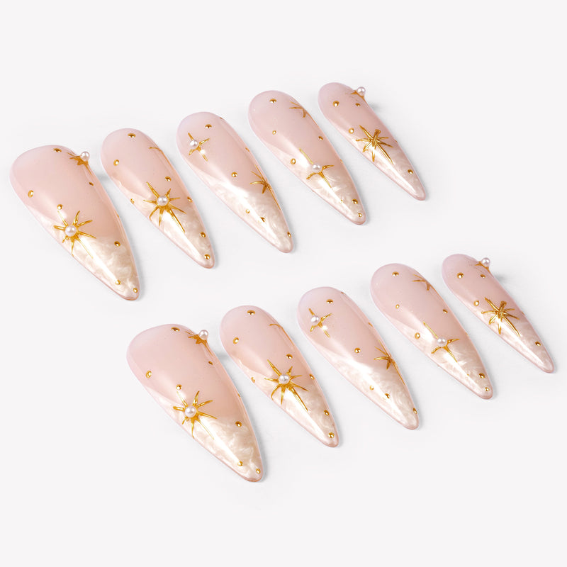 Angled flat lay of Starlit Veil press-on nails with sheer pink bases, white misty tips, gold starbursts, tiny gold studs, and pearl accents