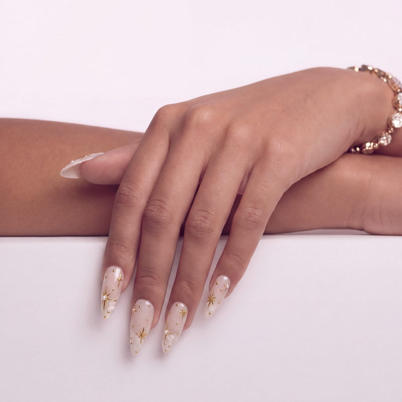 Hand model wearing Starlit Veil press-on nails with sheer pink bases, white misty tips, gold starbursts, gold studs, and pearl accents
