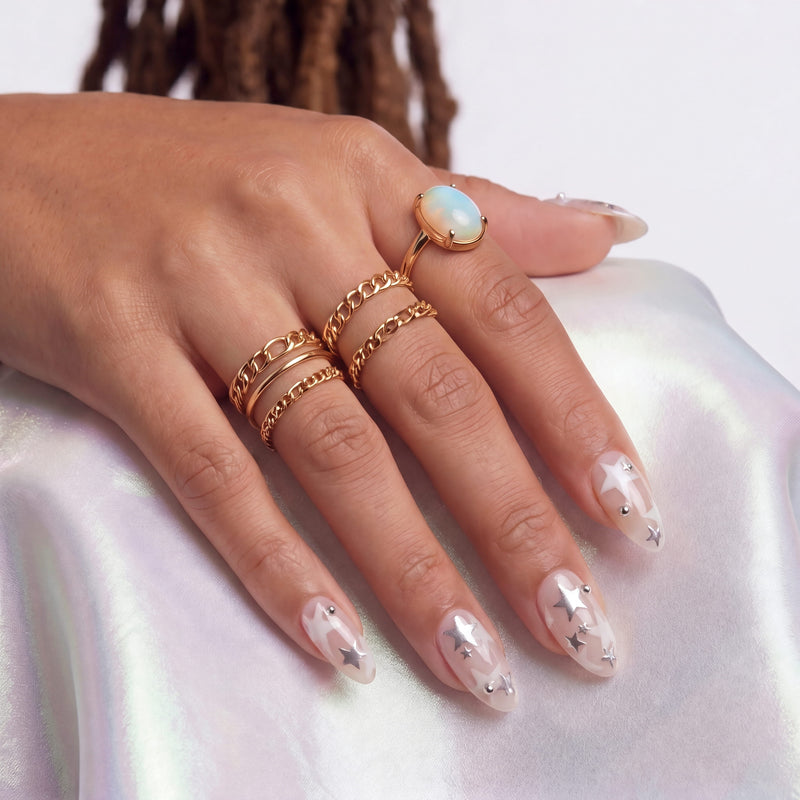 Hand model wearing Starlit Whisper press-on nails with translucent nude pink bases, white star patterns, silver star accents, silver beads, and gold rings on satin fabric