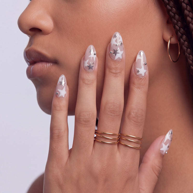 Side face close-up hand model wearing Starlit Whisper press-on nails with translucent nude pink bases, white star patterns, silver star accents, and silver beads