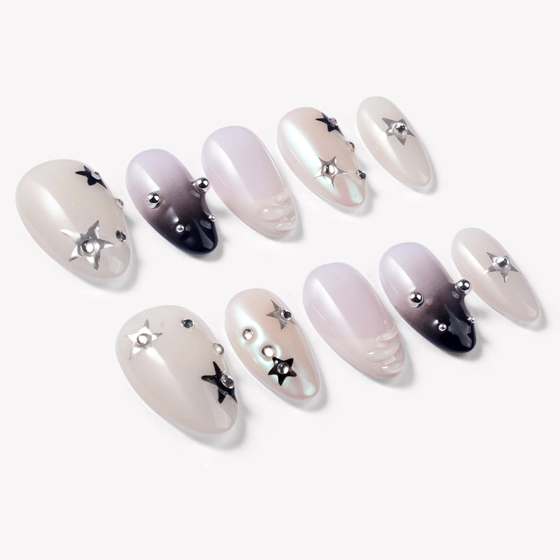 Flat lay of Stellar Muse press-on nails with black and silver star charms, rhinestones, silver beads, glossy nude pink bases, and black gradient accents