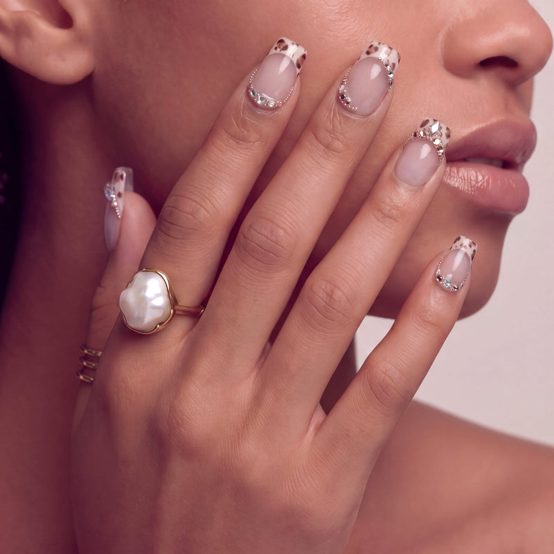 Hand model wearing Sugar Kitten press-on nails with nude pink bases, leopard French tips, and clear rhinestone accents near the face