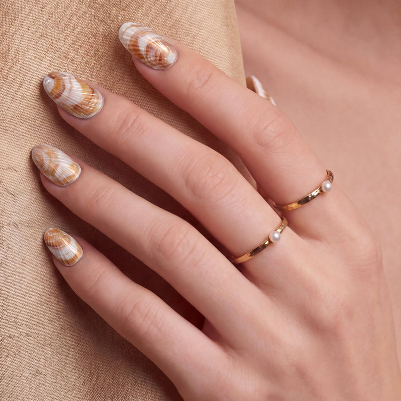 Close-up of a hand wearing brown and white shell-pattern press-on nails with gold rings on a beige background.