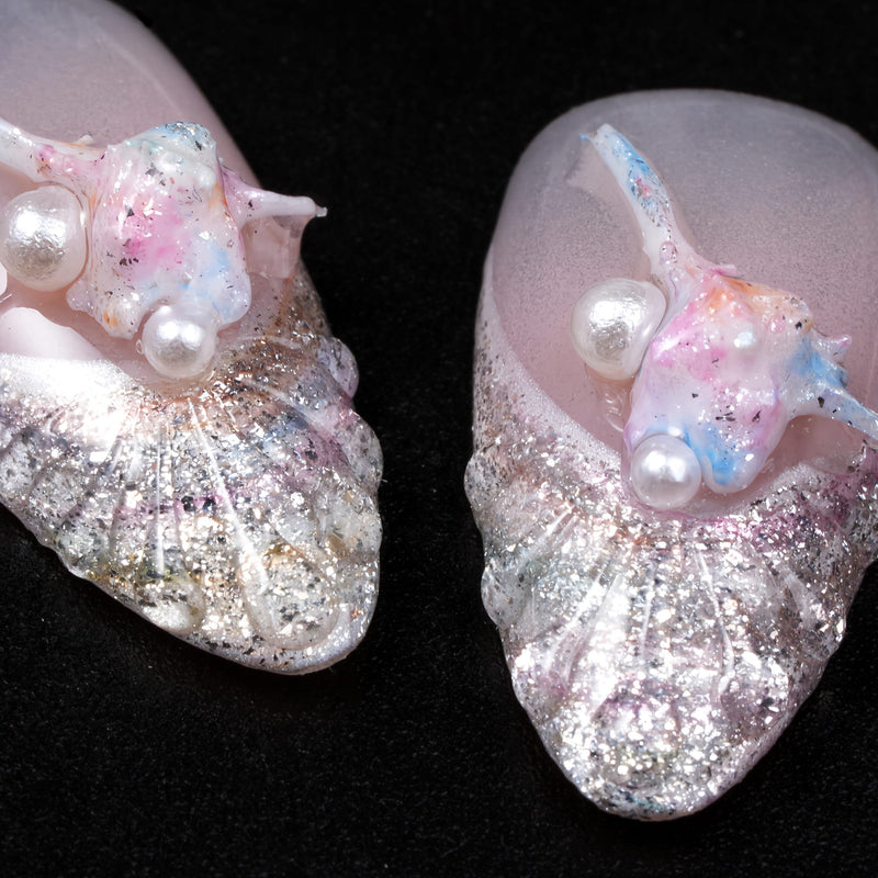 Close-up of two silver shell-inspired press-on nails with pearl and pastel pink-blue accents on a black background.
