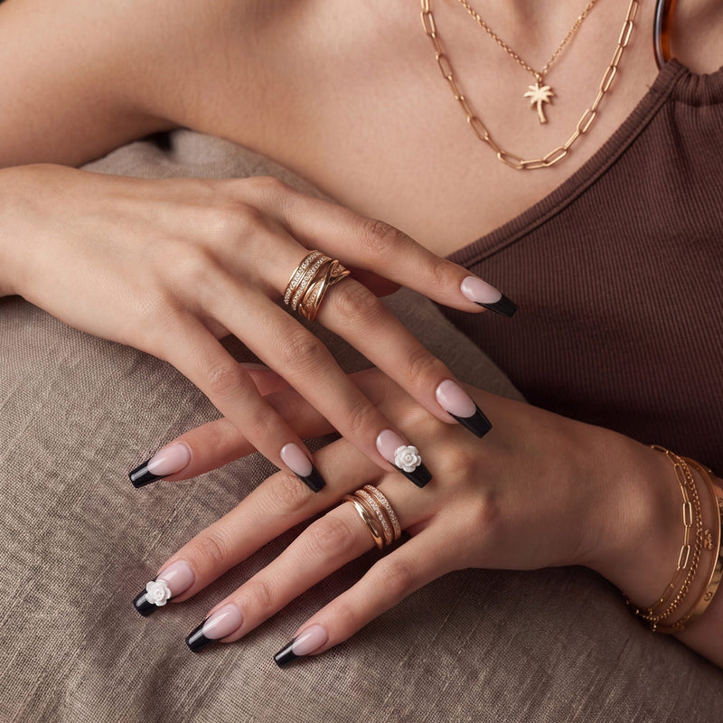 Hand model wearing Timeless Serenity press-on nails with nude bases, black French tips, white 3D flowers, and gold jewelry against a brown top