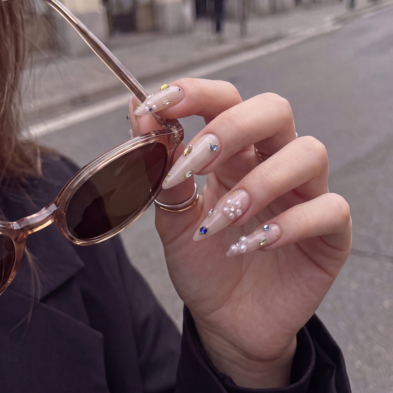 Lifestyle hand model wearing Water Drop Shimmer press-on nails with nude almond bases, pearl clusters, and colorful gem accents while holding sunglasses