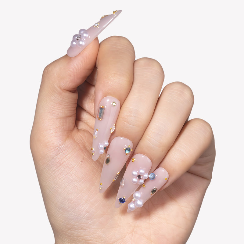 Hand model wearing translucent nude pink stiletto press-on nails with pearl clusters, gold studs, and crystal accents