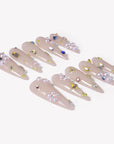 Two rows of translucent nude pink stiletto press-on nails with pearls, gold studs, and crystal accents arranged on a white background