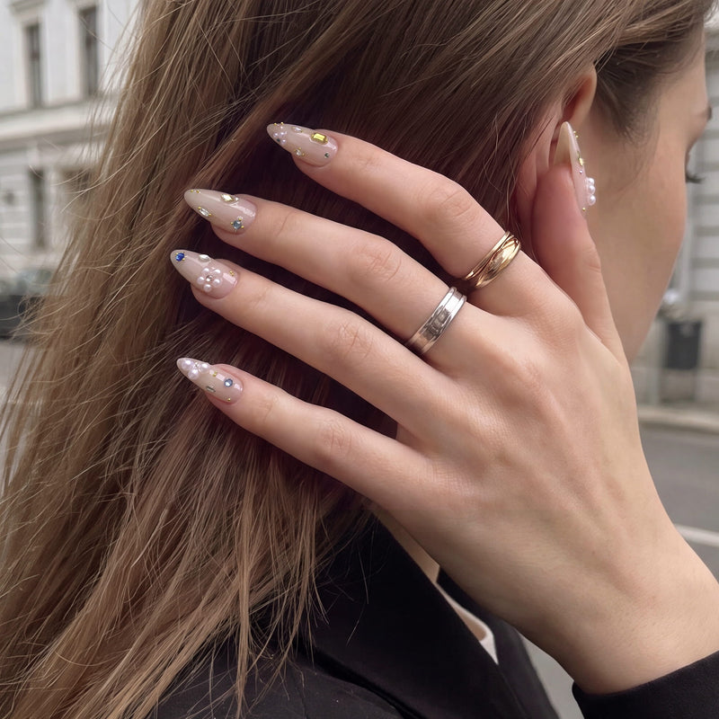 Side hand model wearing Water Drop Shimmer press-on nails with nude almond bases, pearl clusters, colorful gems, and gold bead accents