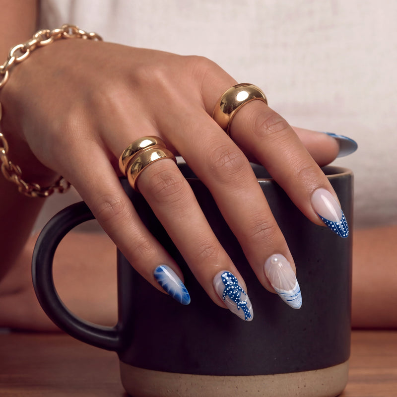 Hand model holding a mug while wearing Whale Song press-on nails with blue ocean wave tips, whale tail design, seashell texture, white dot accents, and pearl details