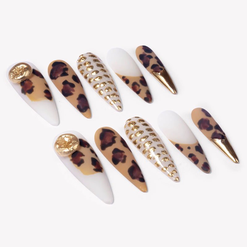Angled flat lay of Wild Majesty press-on nails with tan leopard print, white bases, raised gold dot accents, gold charms, and metallic gold stiletto tips