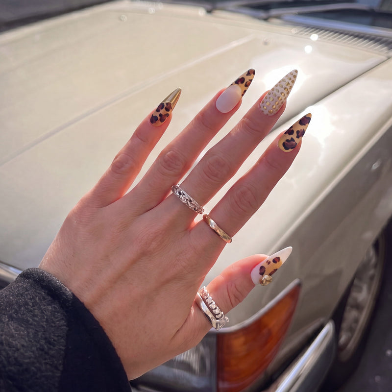 Lifestyle hand model wearing Wild Majesty press-on nails with leopard print, white accent nails, raised gold dots, gold charms, and metallic tips near a car