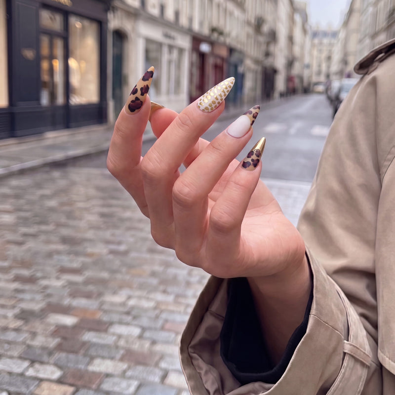 Street hand model wearing Wild Majesty press-on nails with tan leopard print, white accent nails, raised gold dots, and metallic gold stiletto tips