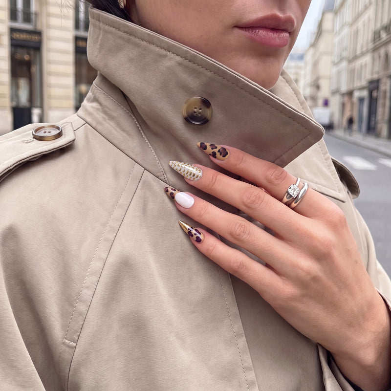 Street style hand model wearing Wild Majesty press-on nails with leopard print, white accents, raised gold dots, and metallic gold stiletto tips against a beige trench coat