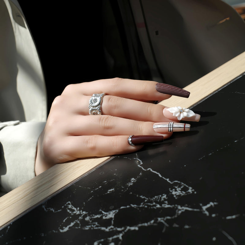 Hand model wearing Winter Serenade press-on nails with purple, plaid, and white floral designs resting on a black tabletop with wood trim