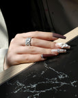 Hand model wearing Winter Serenade press-on nails with purple, plaid, and white floral designs resting on a black tabletop with wood trim